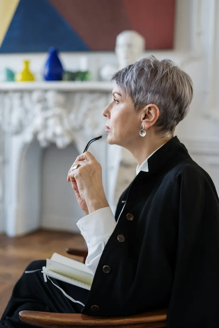 A woman in her fifties pauses mid-thought, reading glasses in hand and a notebook on her lap.