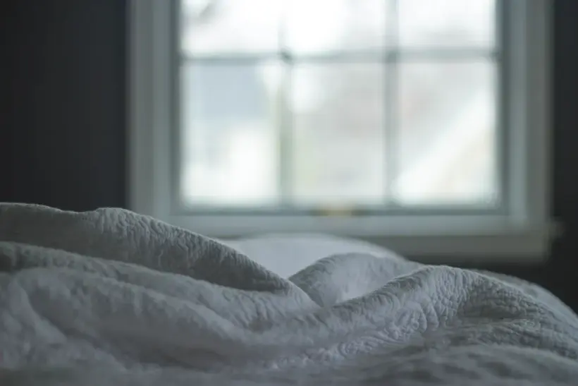An unmade bed with white bedding in front of a bright window.