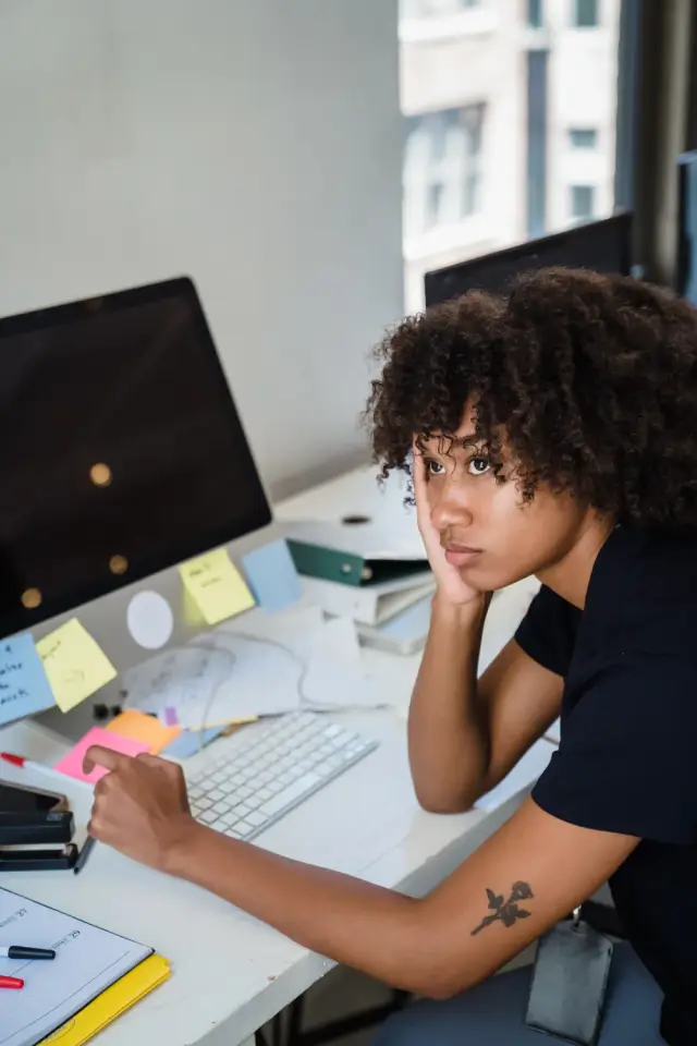 A woman at a cluttered desk covered in sticky notes rests her head on one hand.