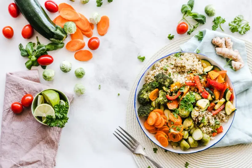 A bowl of quinoa with broccoli, carrots, courgette and peppers, surrounded by fresh vegetables.