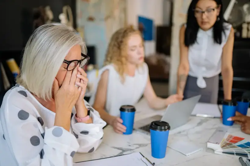 A woman in her fifties presses her fingers to her temple during a work meeting, trying to recall something.