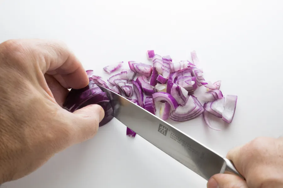 Hands slicing a red onion on a cutting board with a chef's knife.